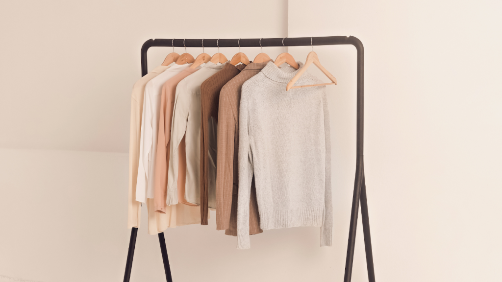 A black clothing rack displays seven neutral-colored sweaters and tops on wooden hangers against a light background, offering inspiration for your next closet cleanout guide.